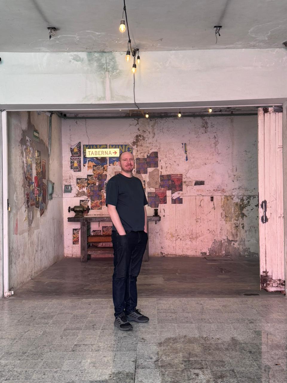 Hrvoje Matosic standing in a Mexico City workshop space — exposed brick walls with peeling concert posters and a 'Taberna' sign behind, hanging pendant bulbs above, a wooden workbench with a vise on the left.