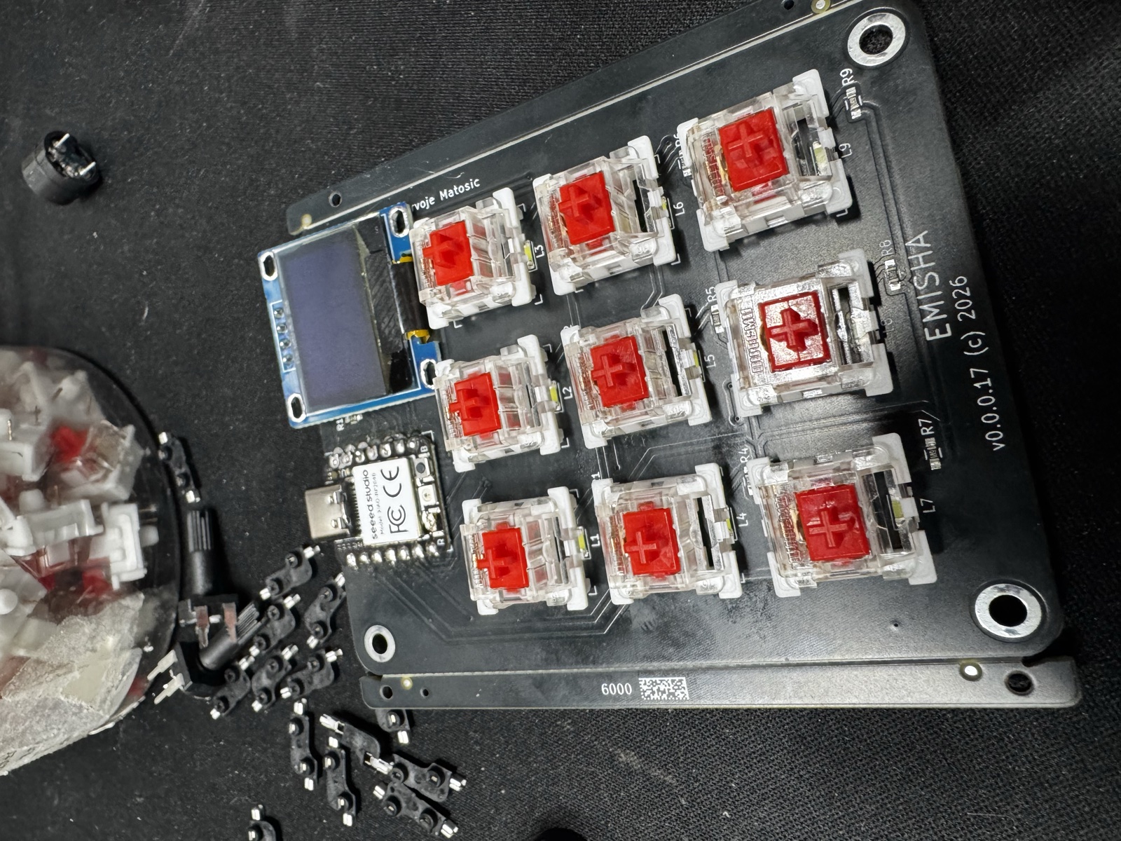 End-of-session workbench. The v0.0.0.17 macropad sits in the centre with all nine red Kailh switches installed in hot-swap sockets. To its left, a small SSD1306 OLED breakout sits loose on the desk, queued up for the next round of experiments.
