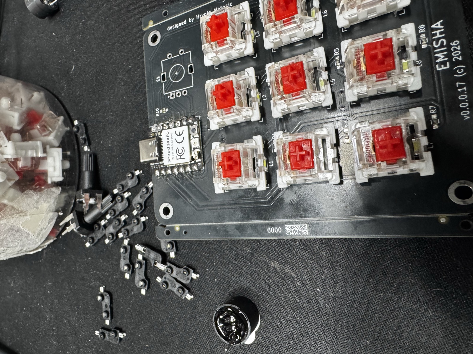 Top-down view of the v0.0.0.17 macropad mid-assembly: XIAO RP2040 flush-soldered at the top edge, eight of nine red Kailh switches seated in hot-swap sockets, scrap tape from the socket strip piled at the bottom-left of the bench.