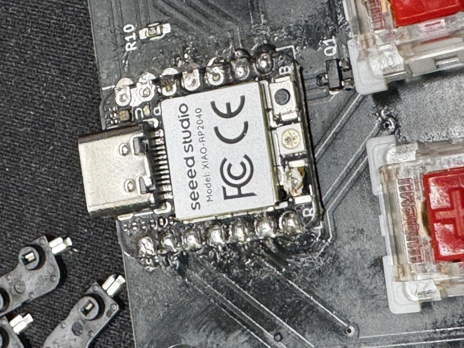 Macro close-up of the XIAO RP2040 flush-soldered to the macropad PCB. The left-side castellation row shows large convex solder blobs that visibly span between adjacent pads, while the right side has smaller distinct fillets. Q1 (an SOT-23 NPN, labelled J3Y) sits intact above the module.